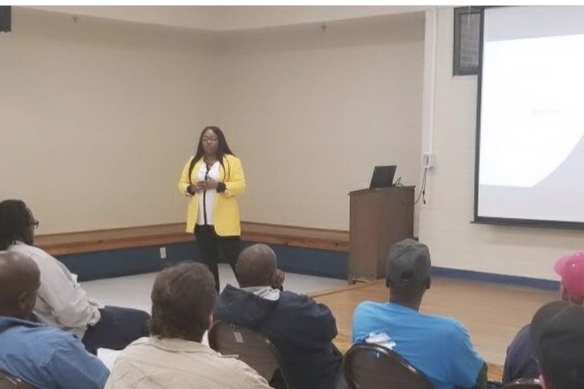 Alyssa Hogan presenting to a seated adult audience in a community hall, a projection screen and lectern beside her