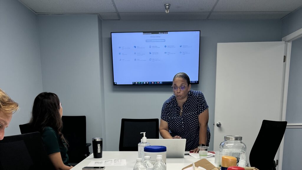 Alyssa Hogan leading a digital literacy training session, participants at the table and a device settings screen displayed behind her