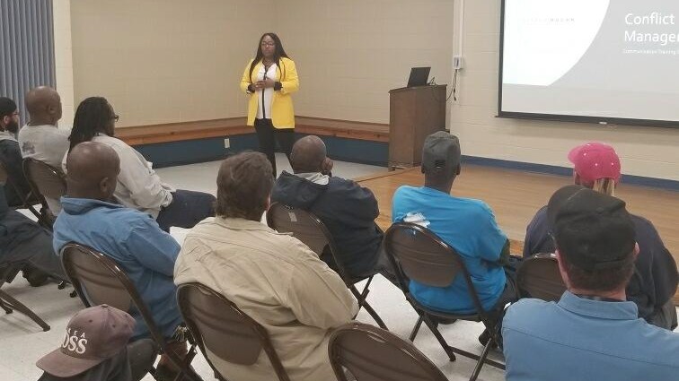 Alyssa Hogan addressing a seated audience at a community workshop, a projected slide behind her