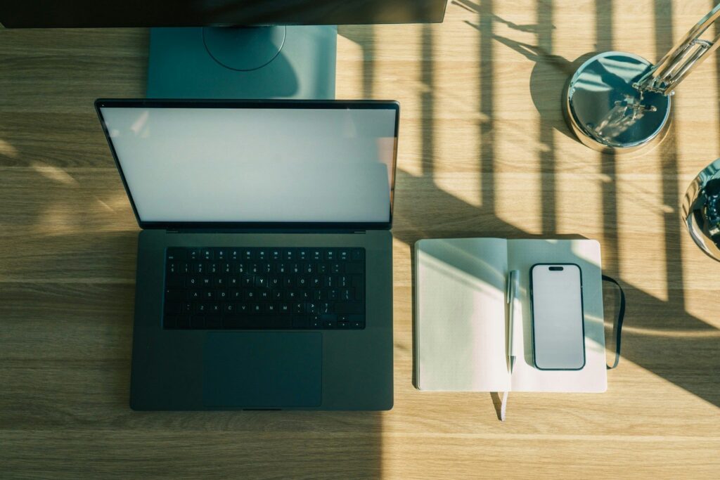 An open laptop, a notebook and a phone on a wooden desk